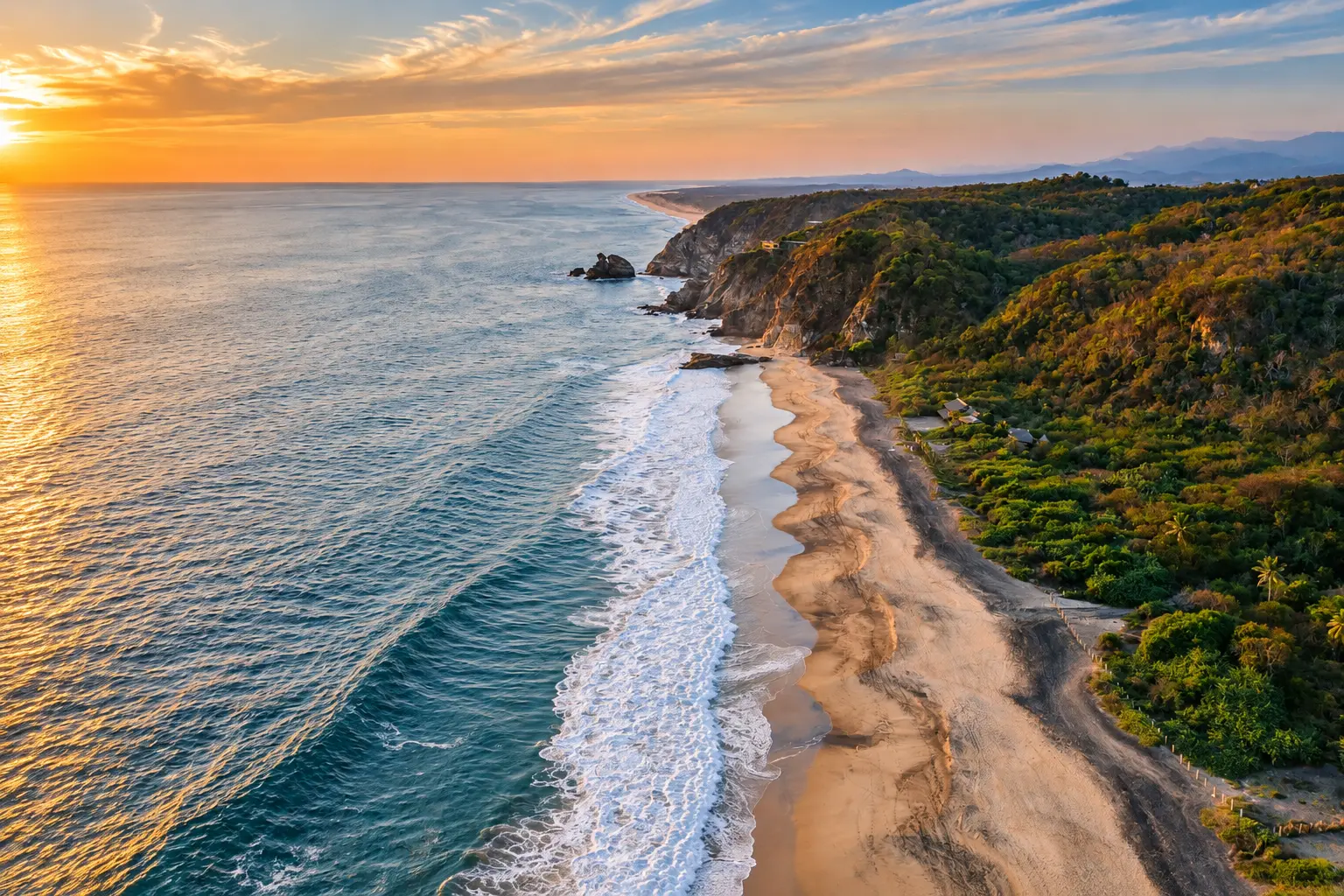 Aerial view of the Pacific coastline at golden hour with waves rolling onto a sandy beach below forested cliffs